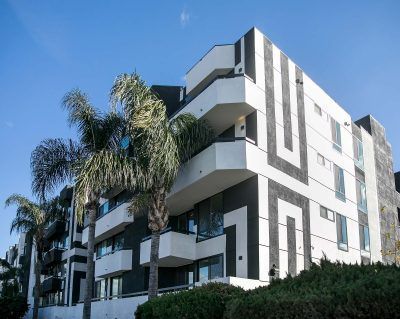 Modern, multi-story apartment building with white and dark gray geometric panels, balconies, and palm trees against blue sky.