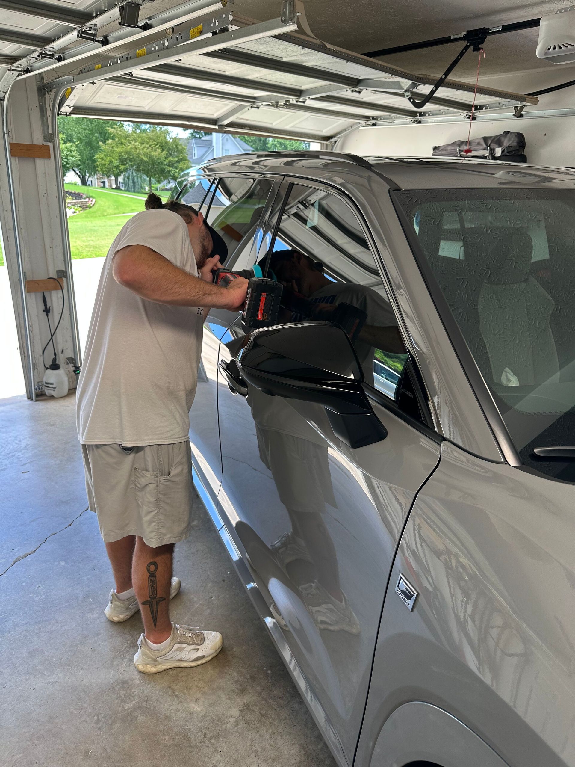 Man working on the window of a light-colored car inside a garage. He is holding a tool and looking closely.