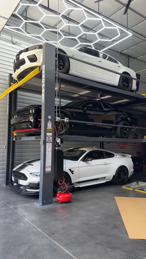 Cars on a multi-level lift in a garage with honeycomb ceiling lights. White and black vehicles.