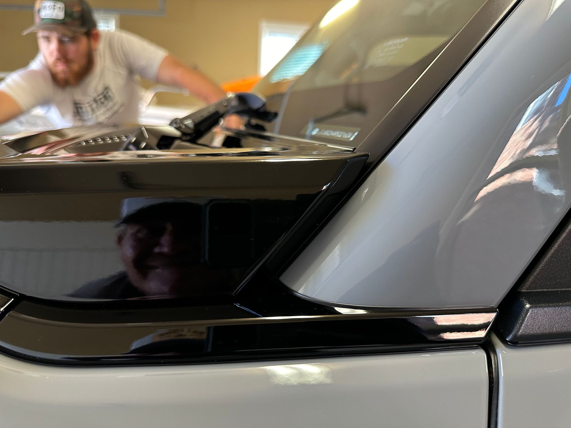 Man working on the hood of a white vehicle; shiny black trim and reflection of the man visible.