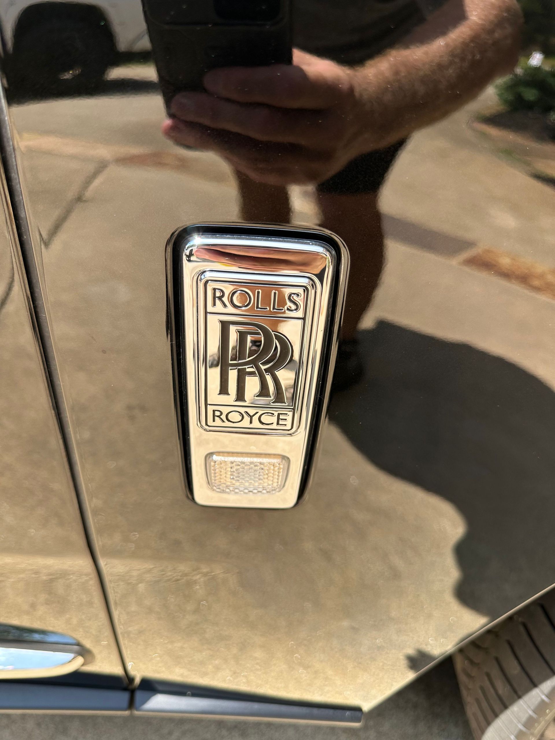 Rolls-Royce logo on a car door, reflecting a person holding a phone. Chrome emblem on black paint.