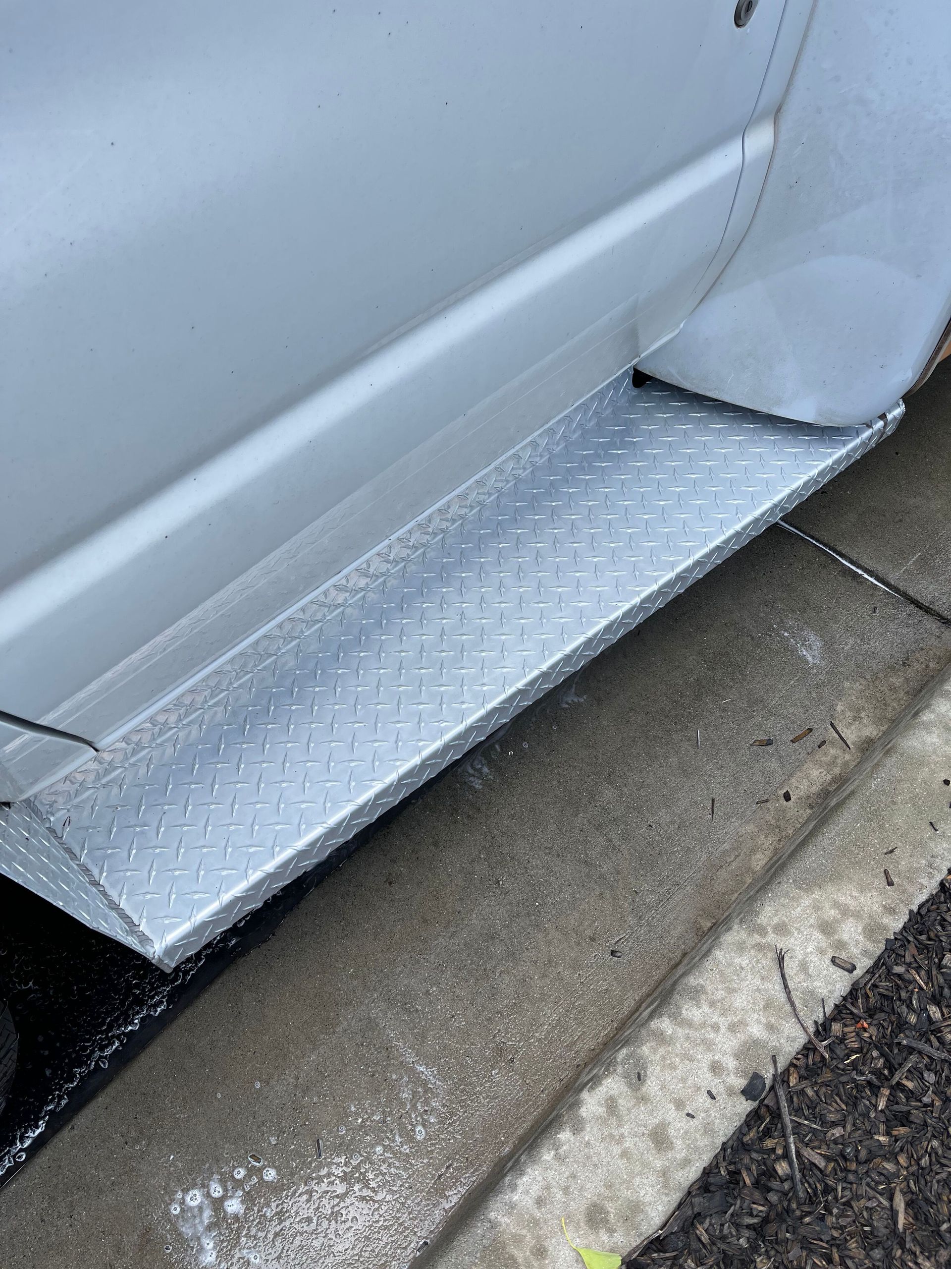 Silver diamond plate step attached to a white vehicle, positioned on a wet concrete surface.