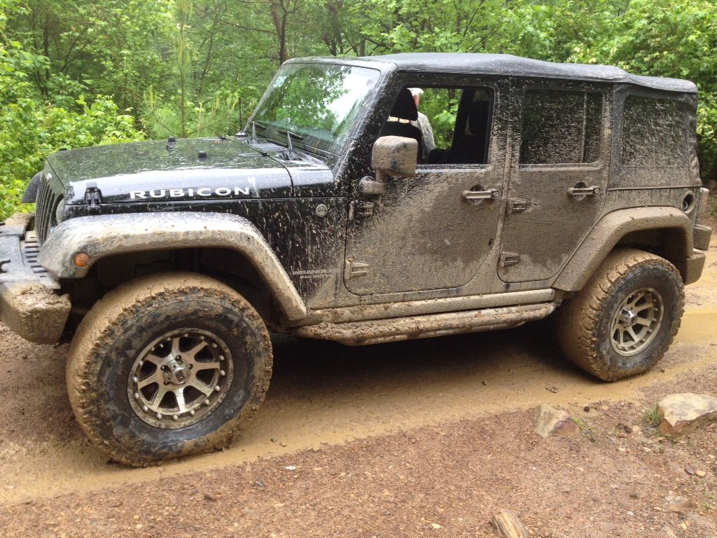 Mud-covered black Jeep Rubicon off-roading on a muddy trail in a wooded area.