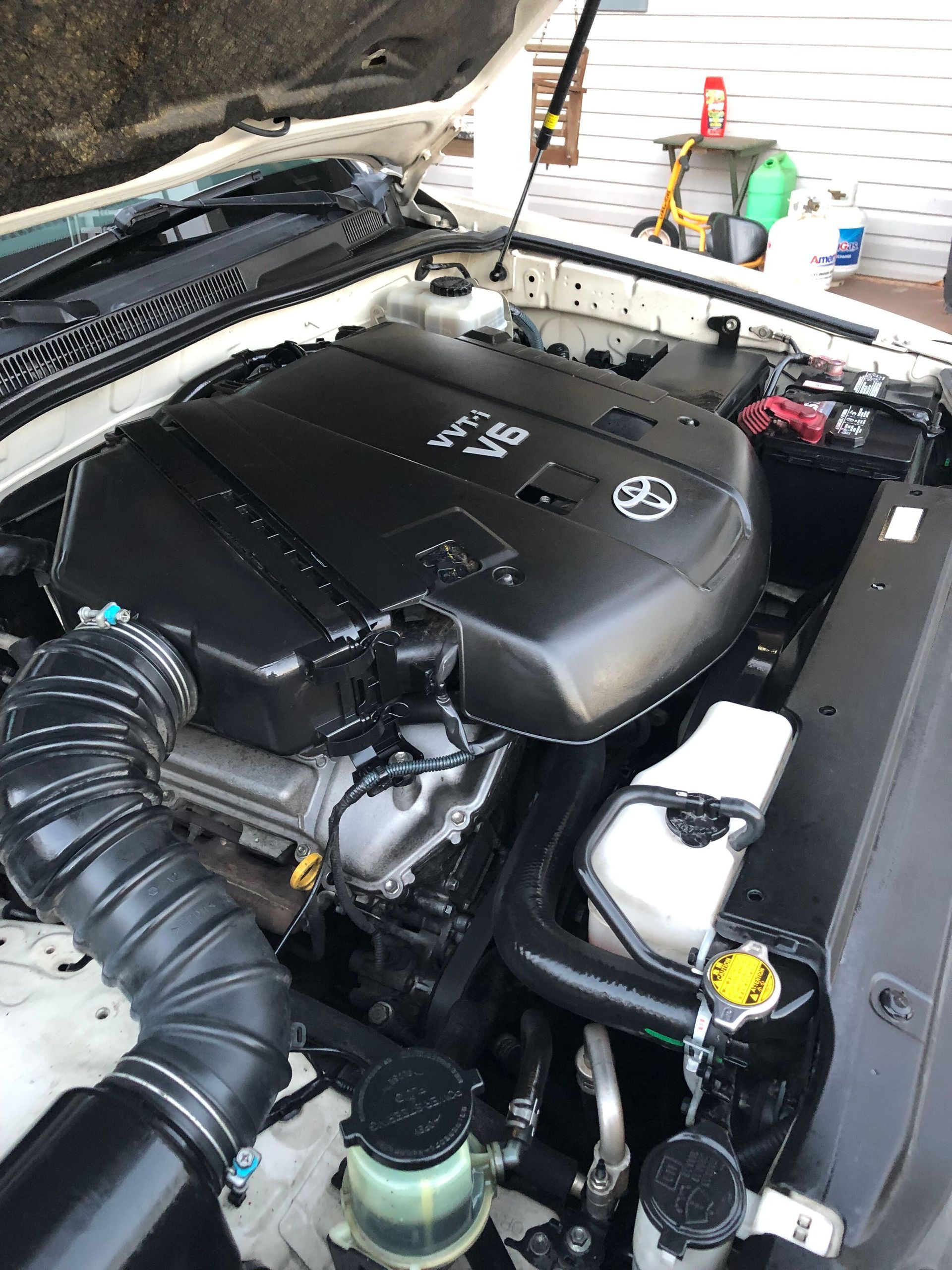 Clean Toyota 4.0L engine bay; black engine cover, white car body, various fluid reservoirs, and air intake.