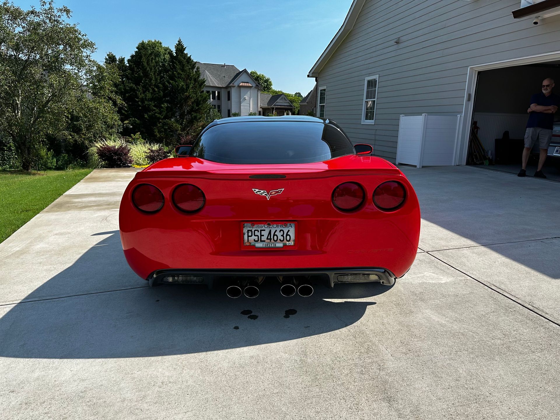 Red Chevrolet Corvette parked on a driveway in front of a house, with a person standing by the open garage.