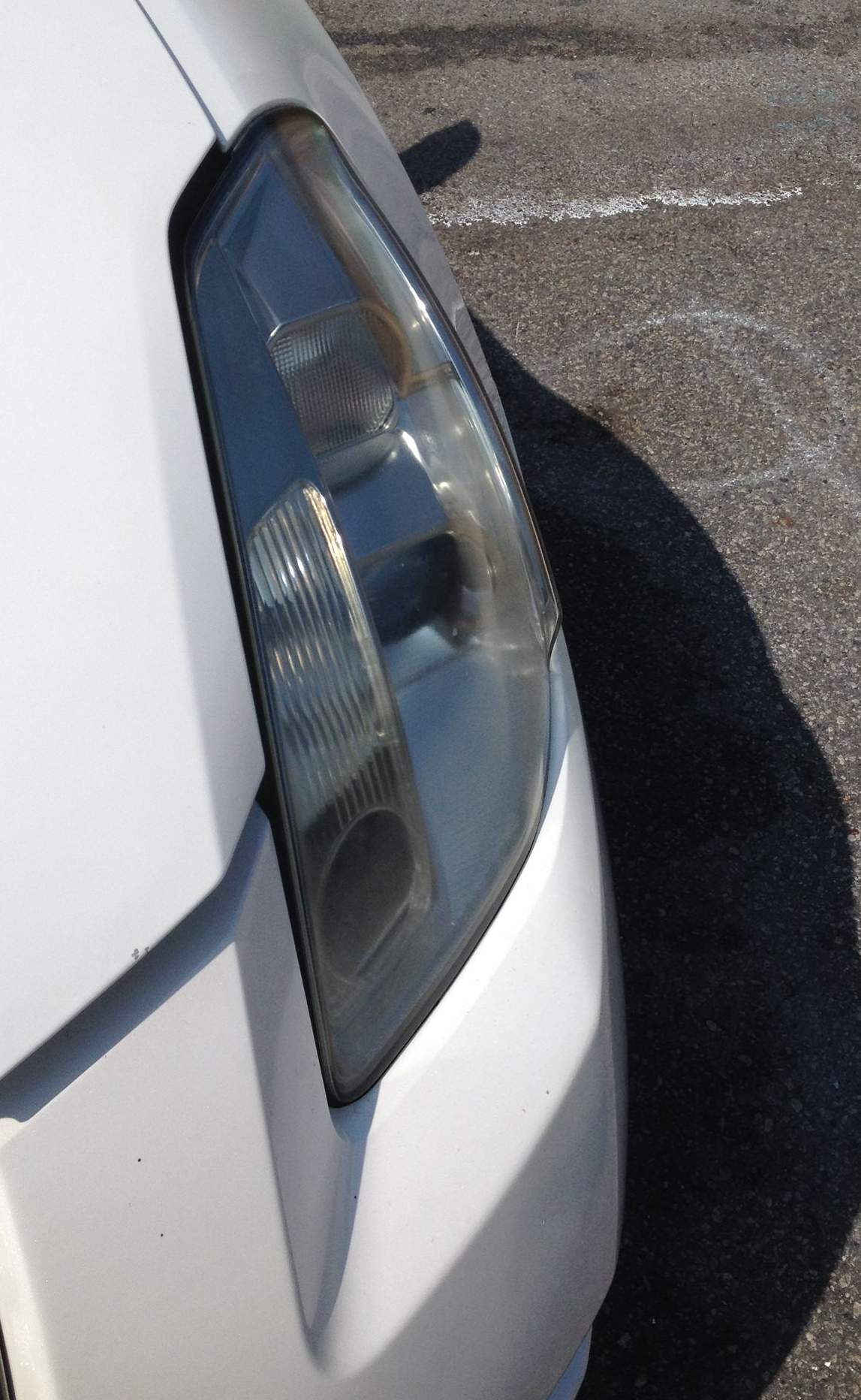 White car's front headlight against asphalt. Sunlight casts a shadow.