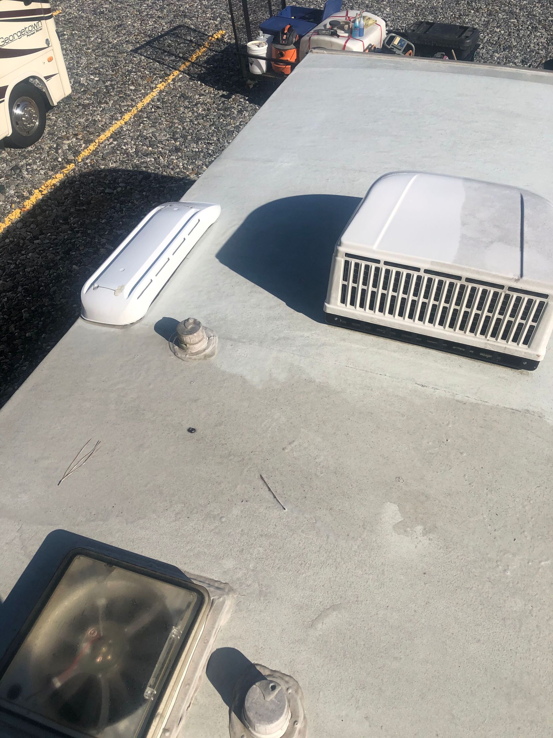 RV roof with a vent, air conditioner, and other fixtures. Gray surface, sunny day.