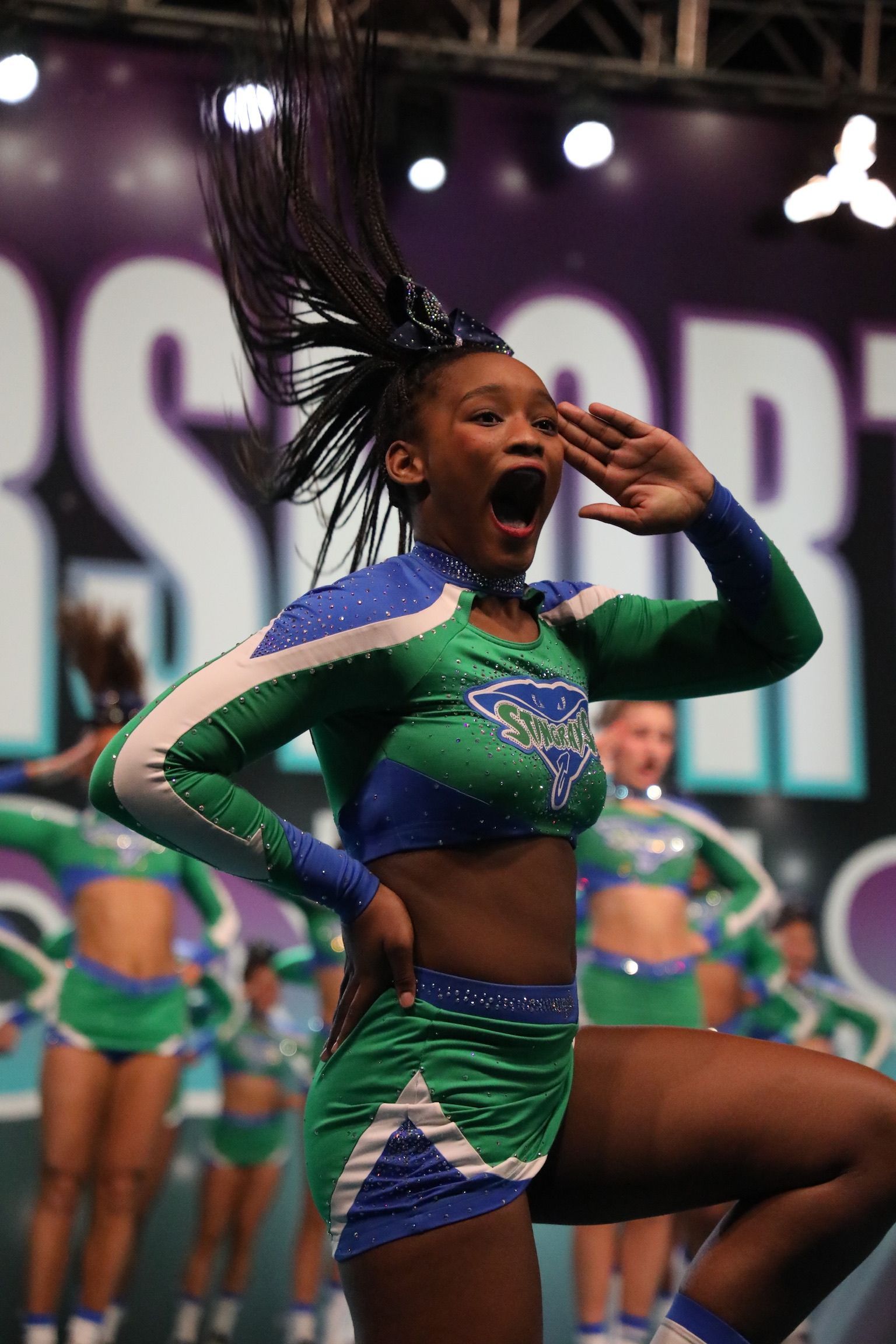 A team of cheerleaders in vibrant green and blue uniforms performs on a stage, with one cheering in the foreground.