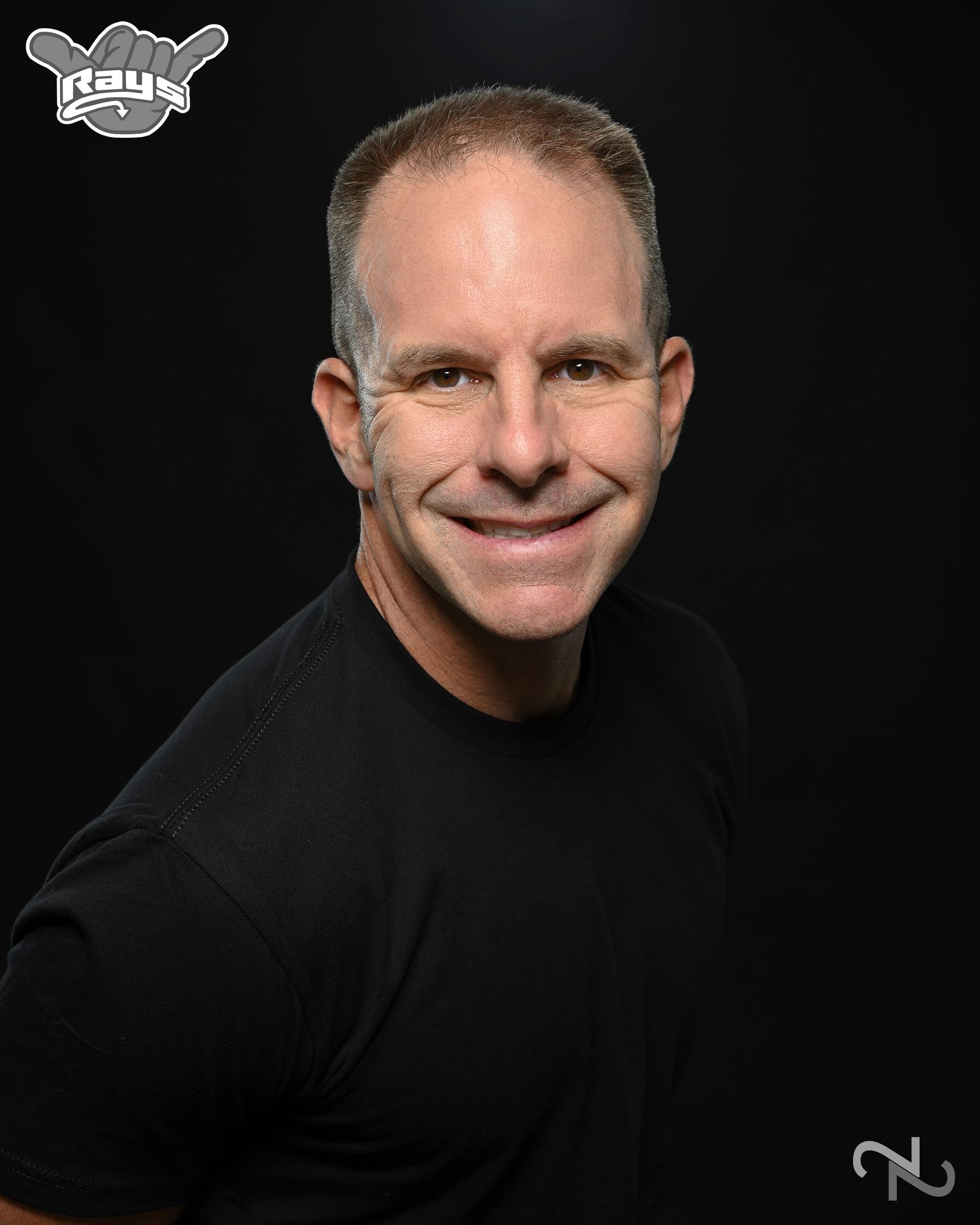A man in a black shirt is smiling in front of a black background.