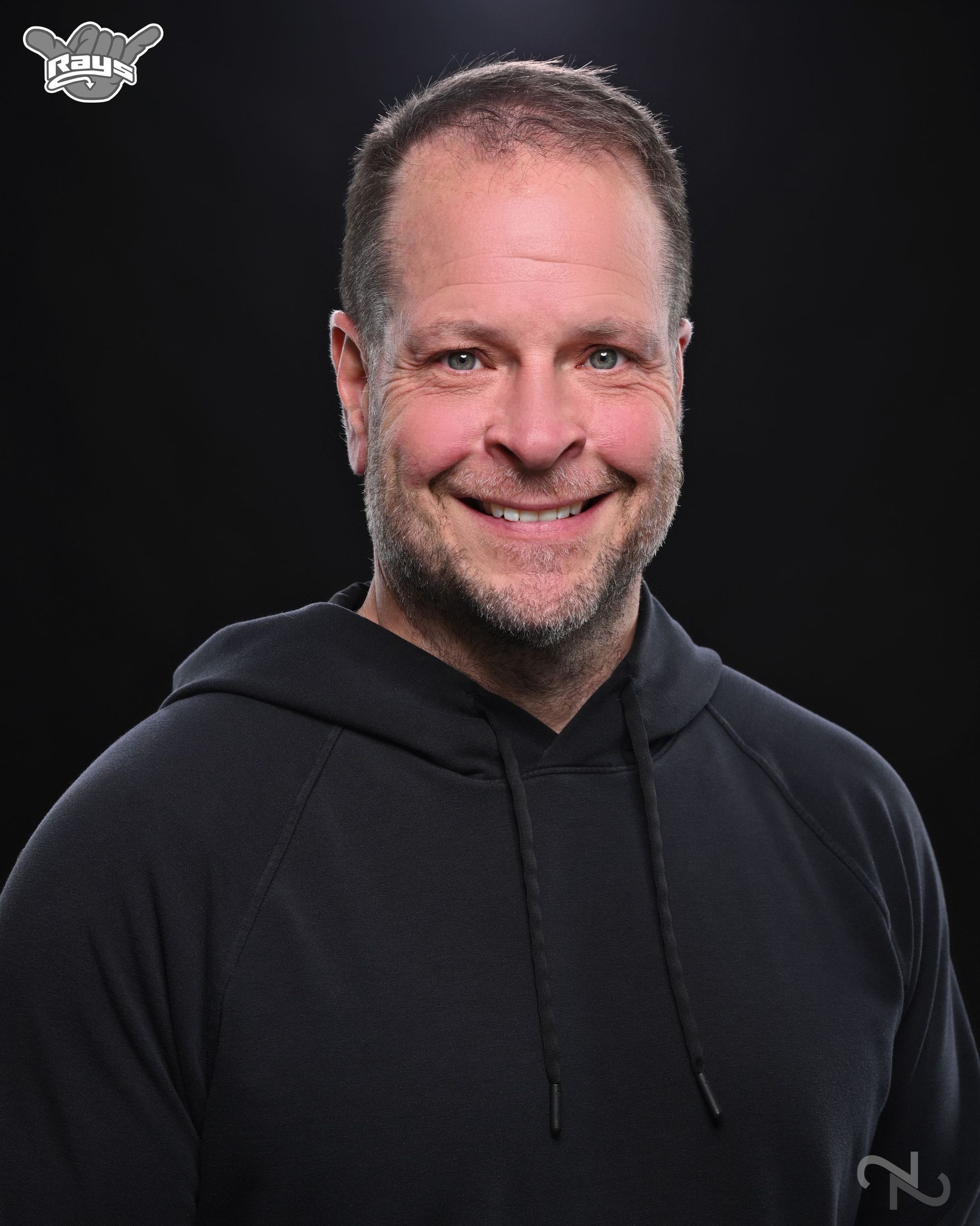 A man wearing a black hoodie is smiling in front of a black background.