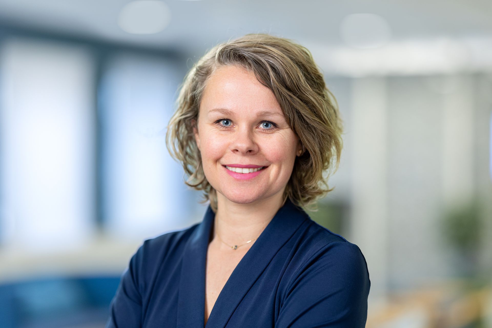 Een vrouw in een blauw shirt lacht naar de camera, met haar armen over elkaar.