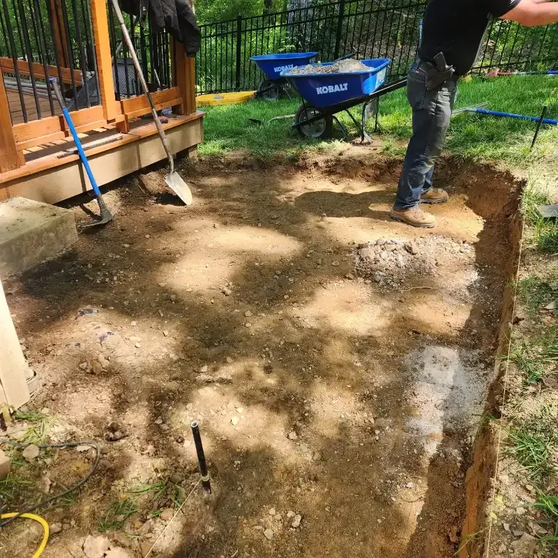A man is digging a hole in the ground in front of a blue wheelbarrow.
