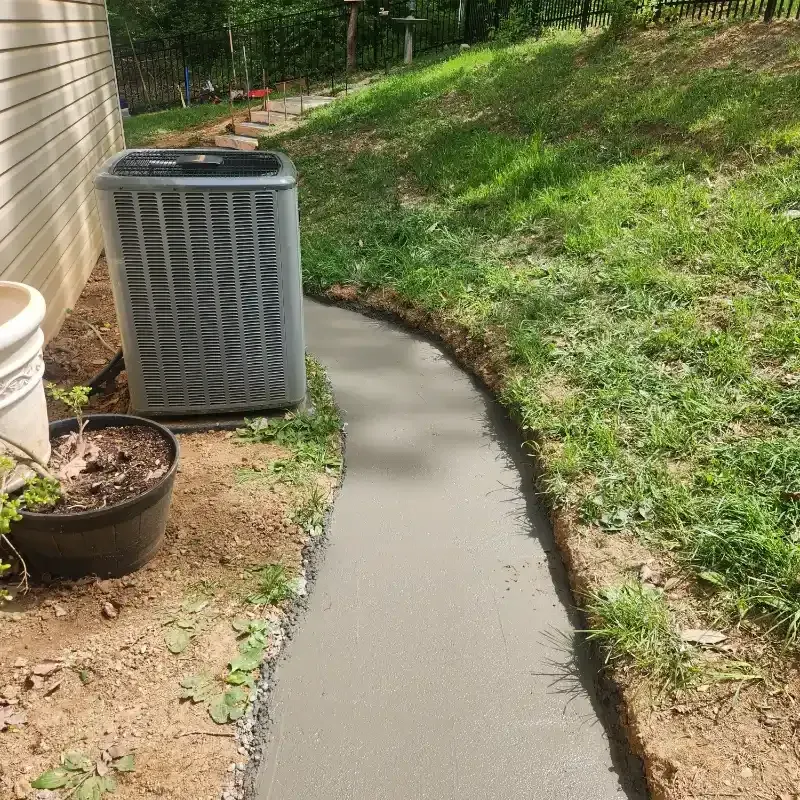 A gray air conditioner is sitting next to a concrete walkway.