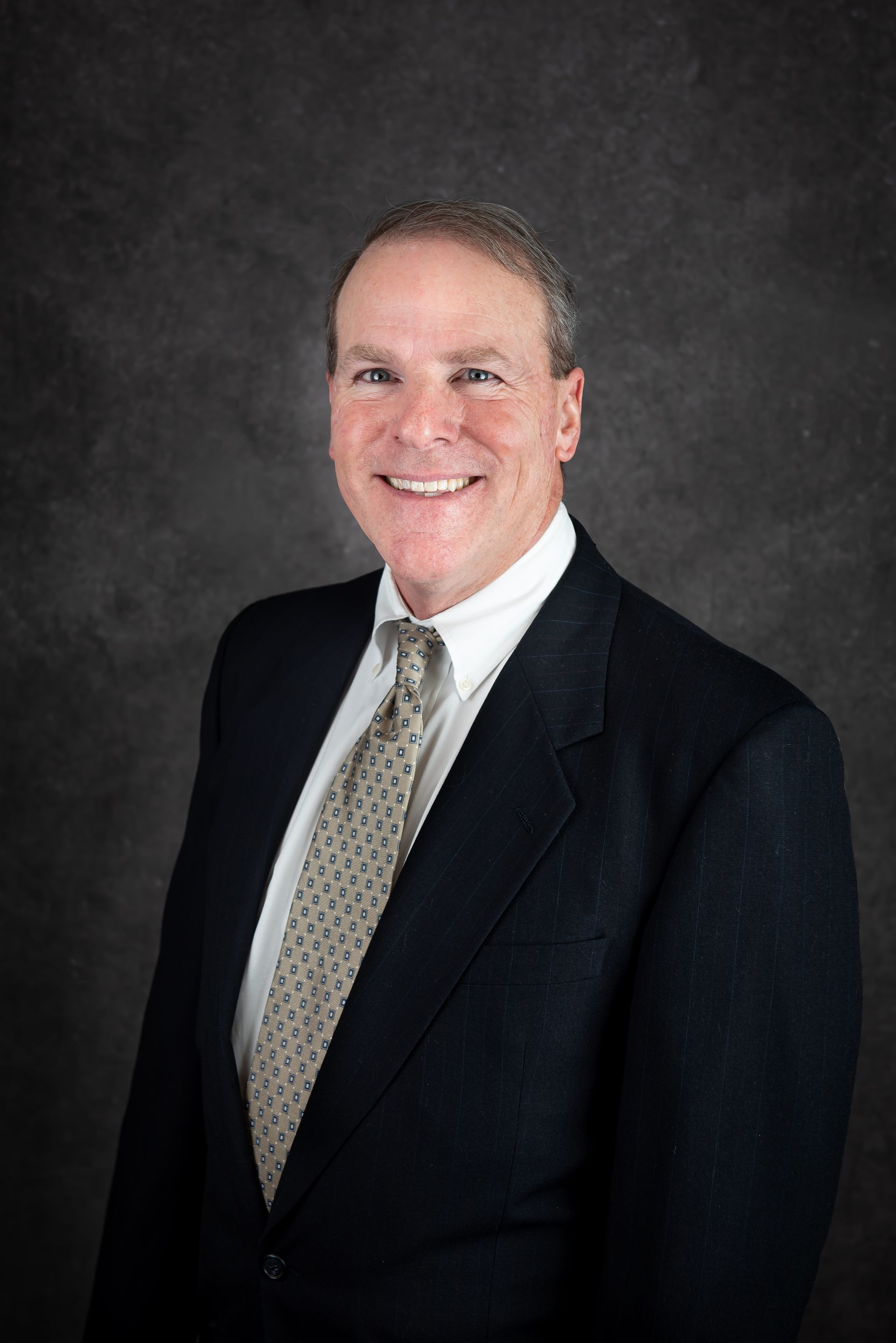 Man in dark suit and tie, smiling, posed against a dark gray backdrop.