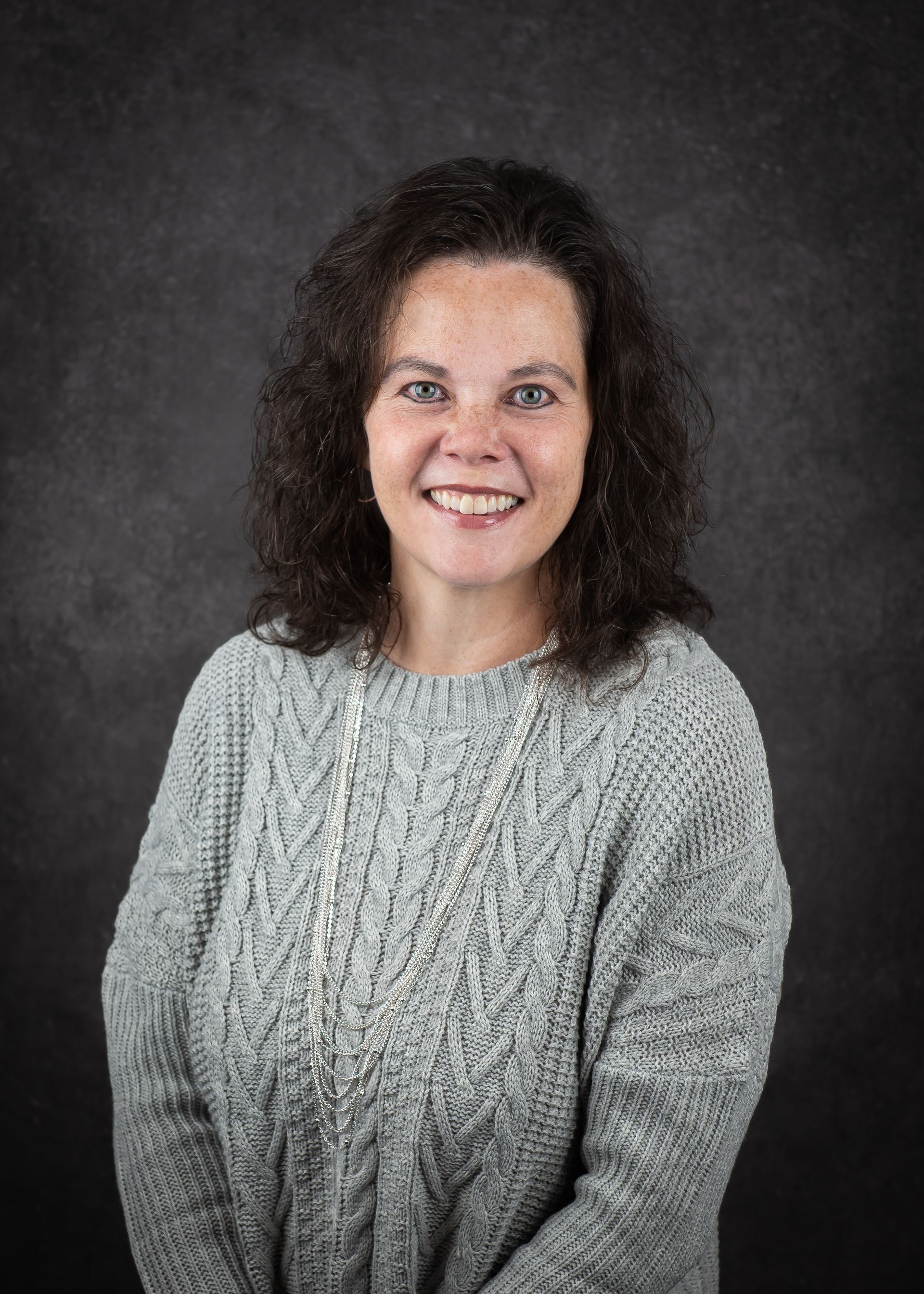 Woman with curly brown hair, smiling, wearing a gray sweater and necklace against a dark background.