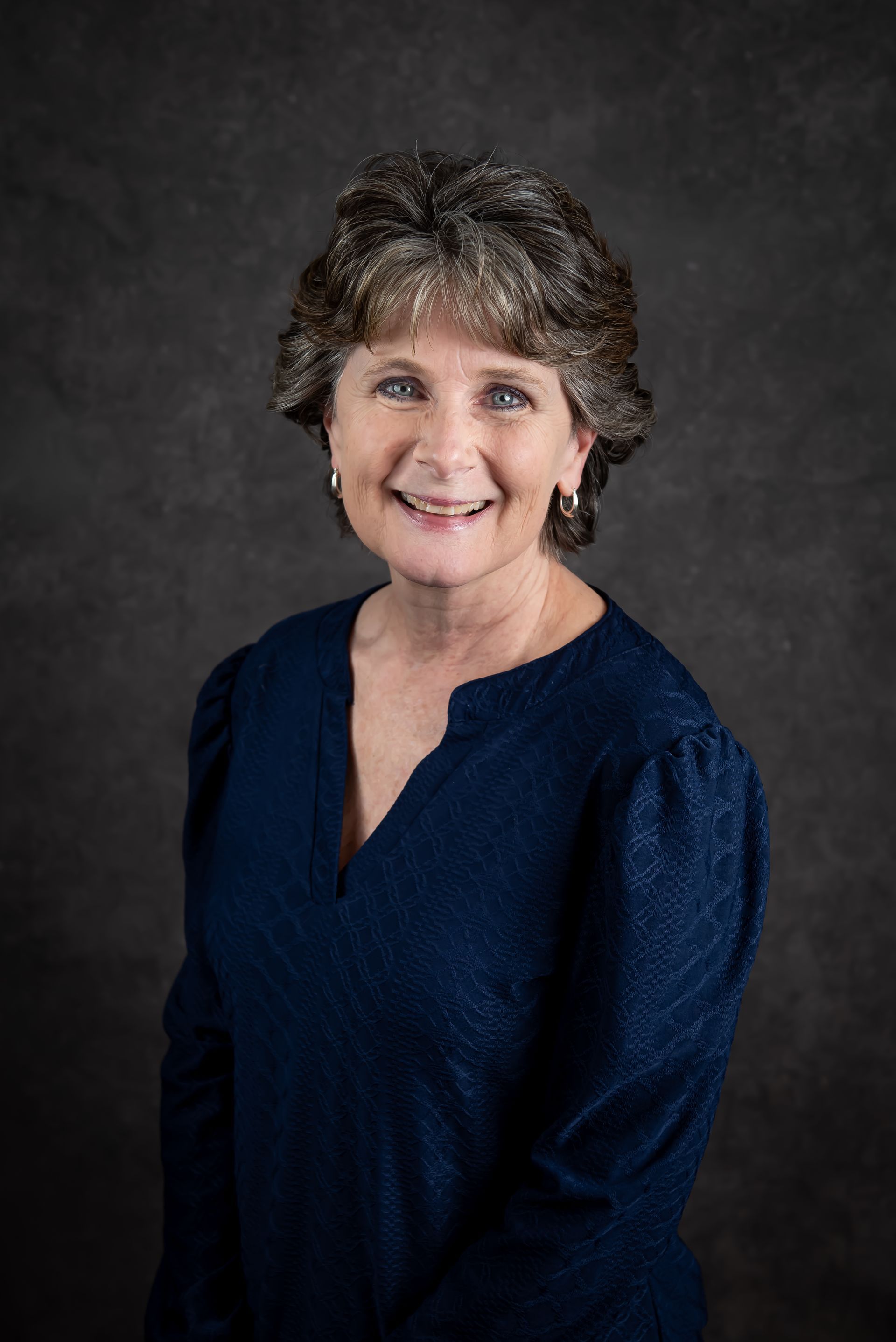 Woman with short brown hair smiles, wearing a navy blue top, against a dark gray background.