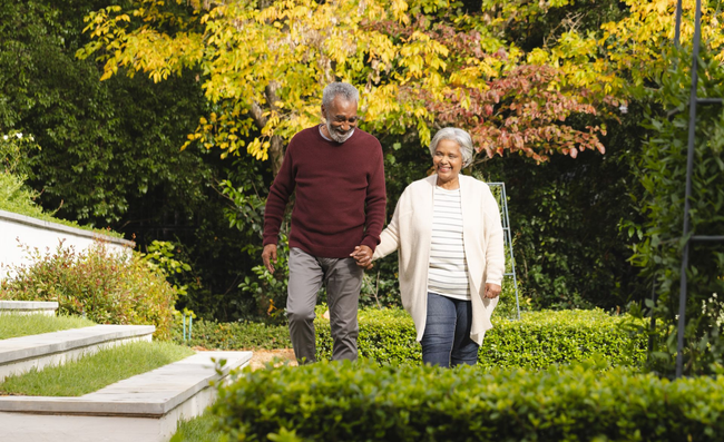 Elderly couple holding hands walking in a garden with green and yellow foliage.