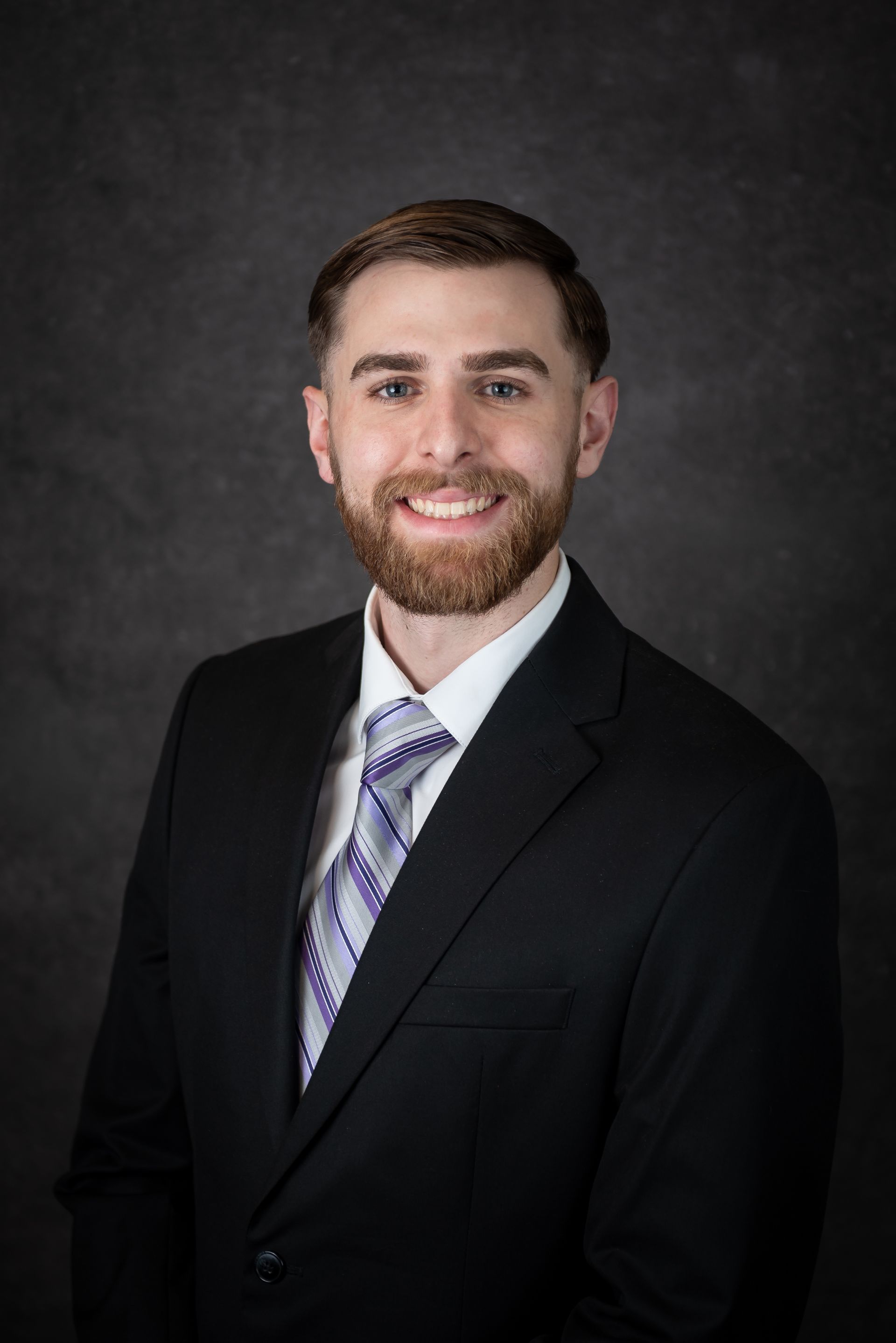 Man in a suit smiles at the camera, with a dark background.