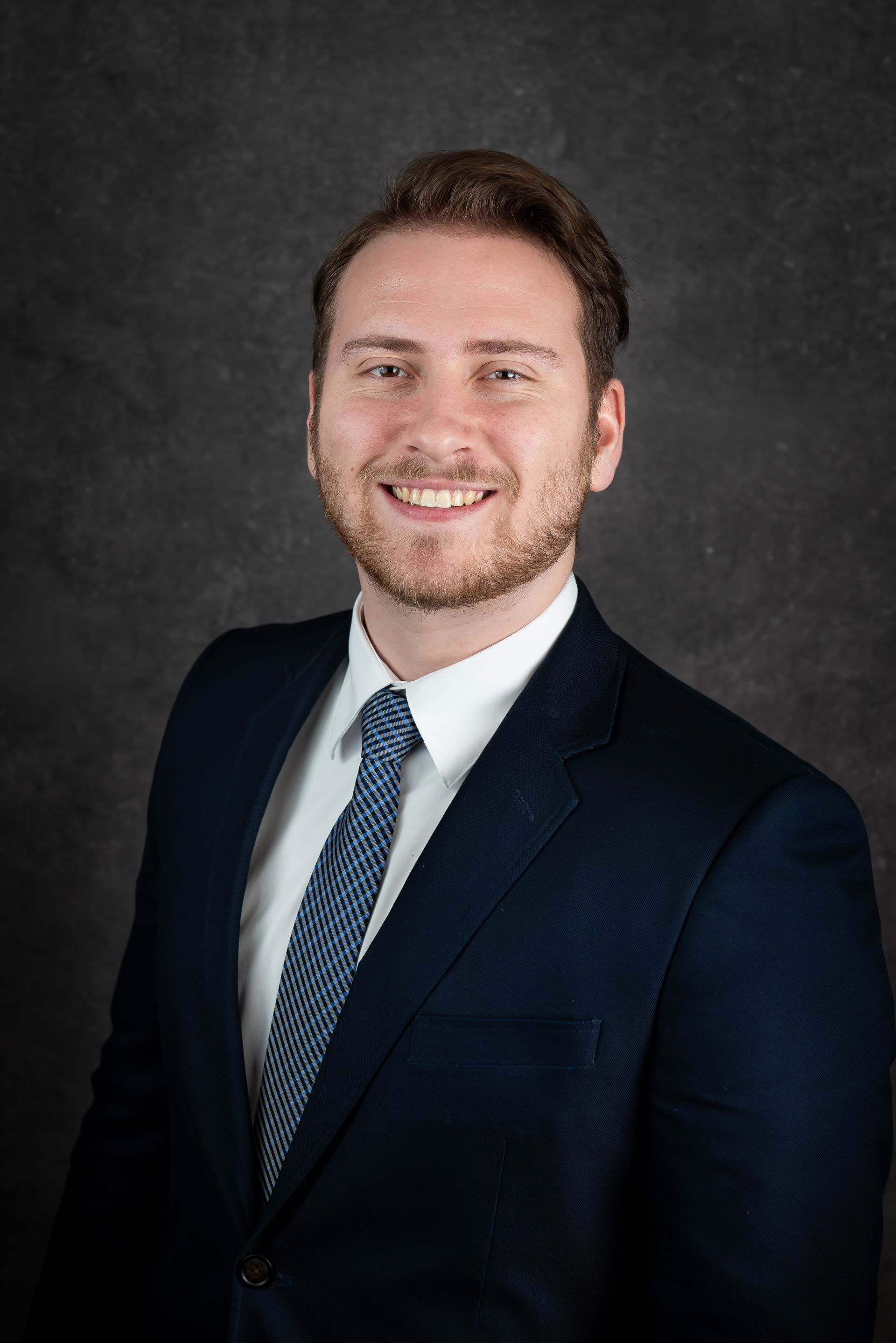 Man in a navy blue suit and tie smiles against a dark gray backdrop.