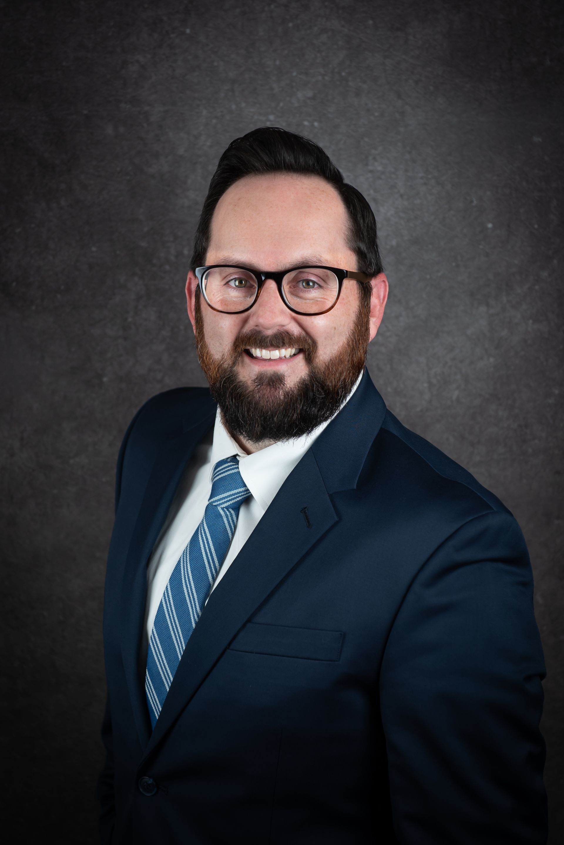 Man with a beard and glasses smiling, wearing a blue suit and tie, against a dark background.