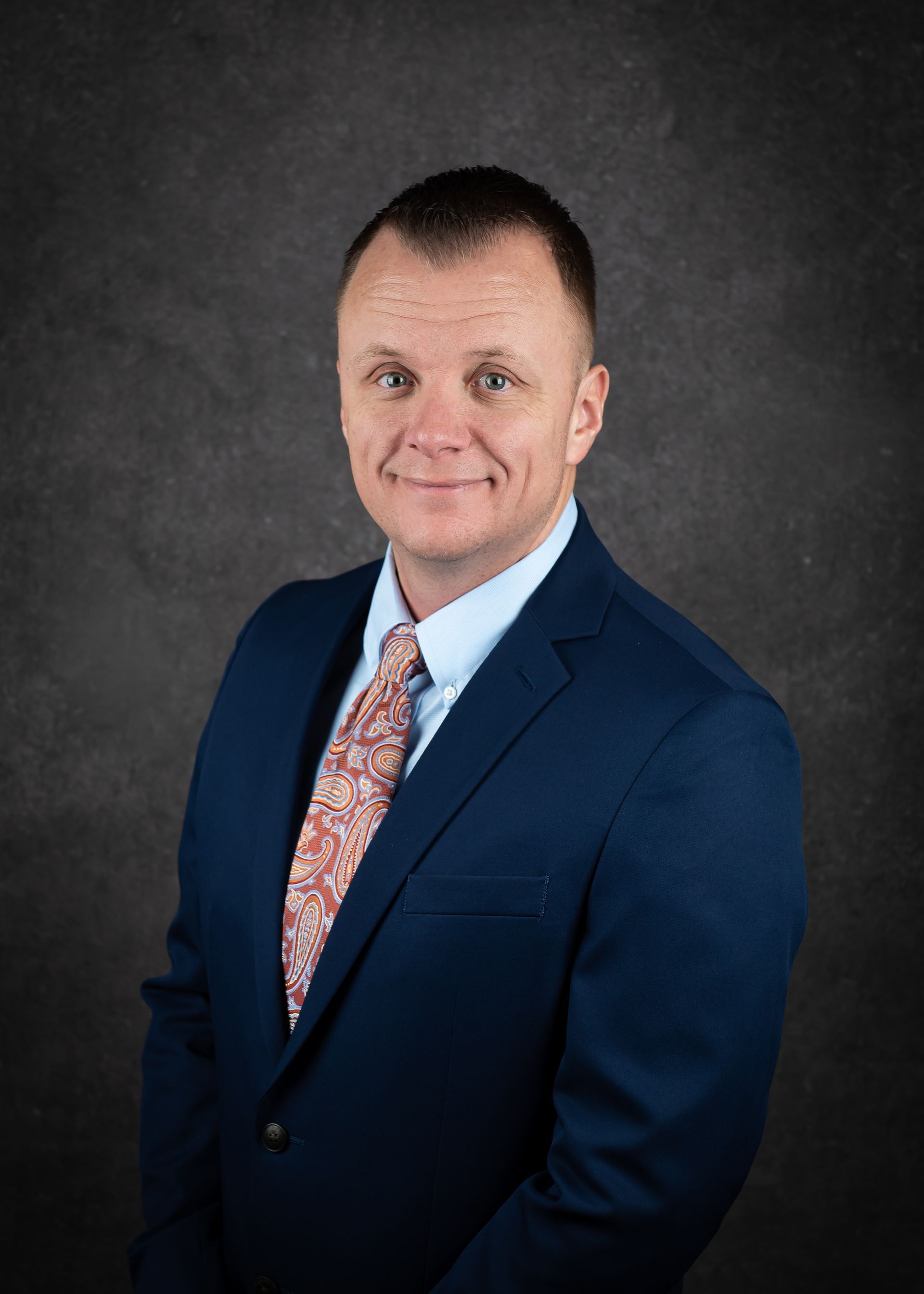 Man in blue suit, patterned tie, light blue shirt, against a dark gray background, smiling.