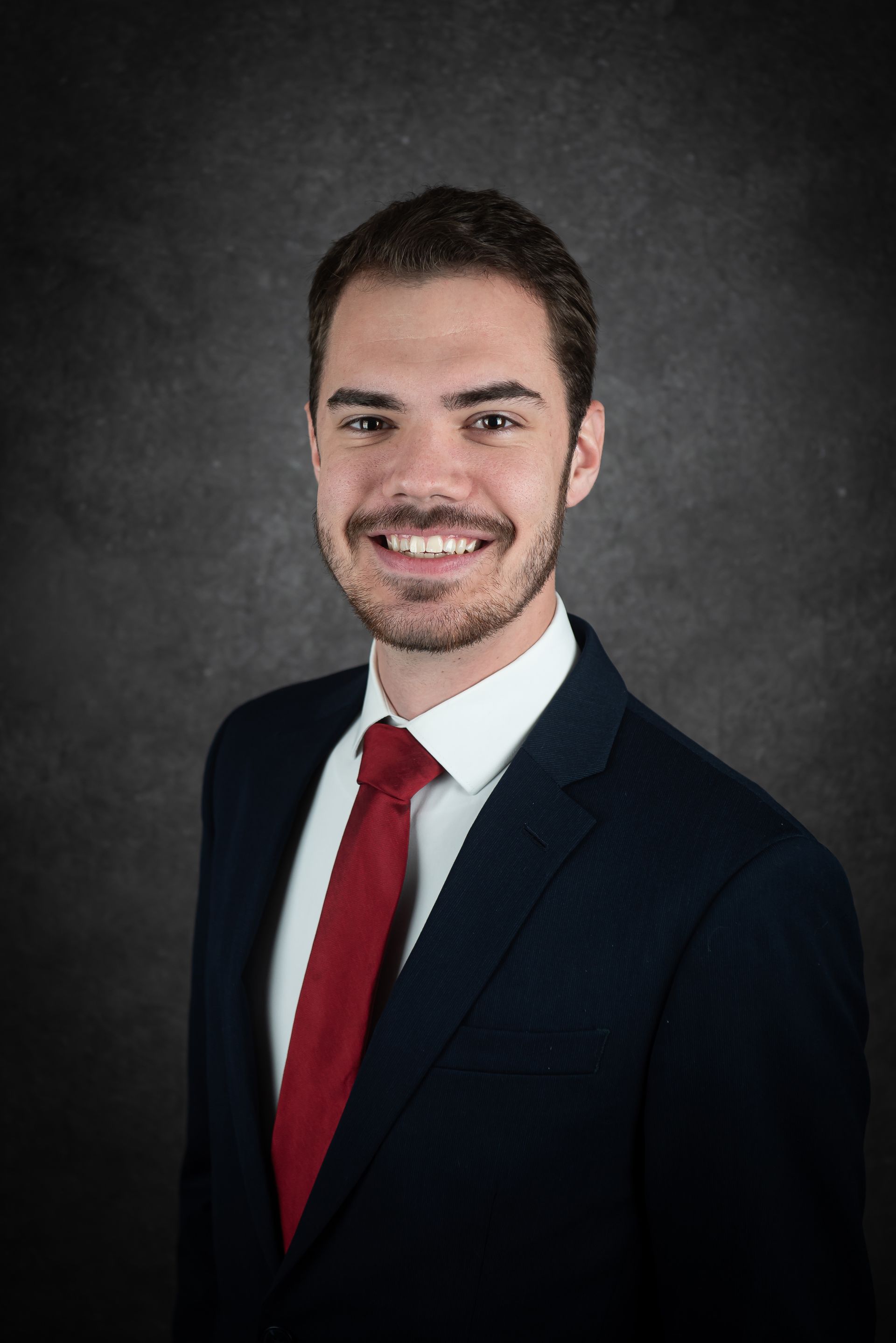 Man in navy suit, white shirt, and red tie smiling, gray backdrop.