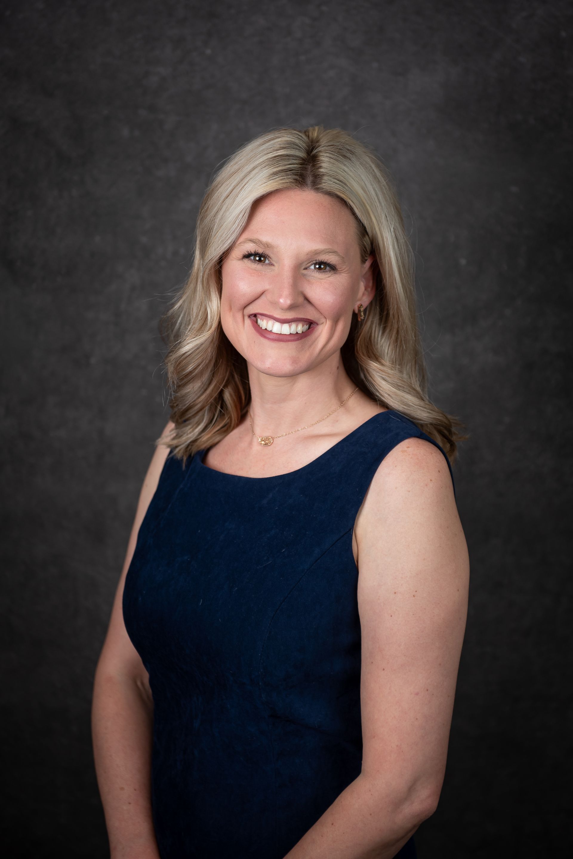 Woman smiling in a dark blue sleeveless dress against a dark background.