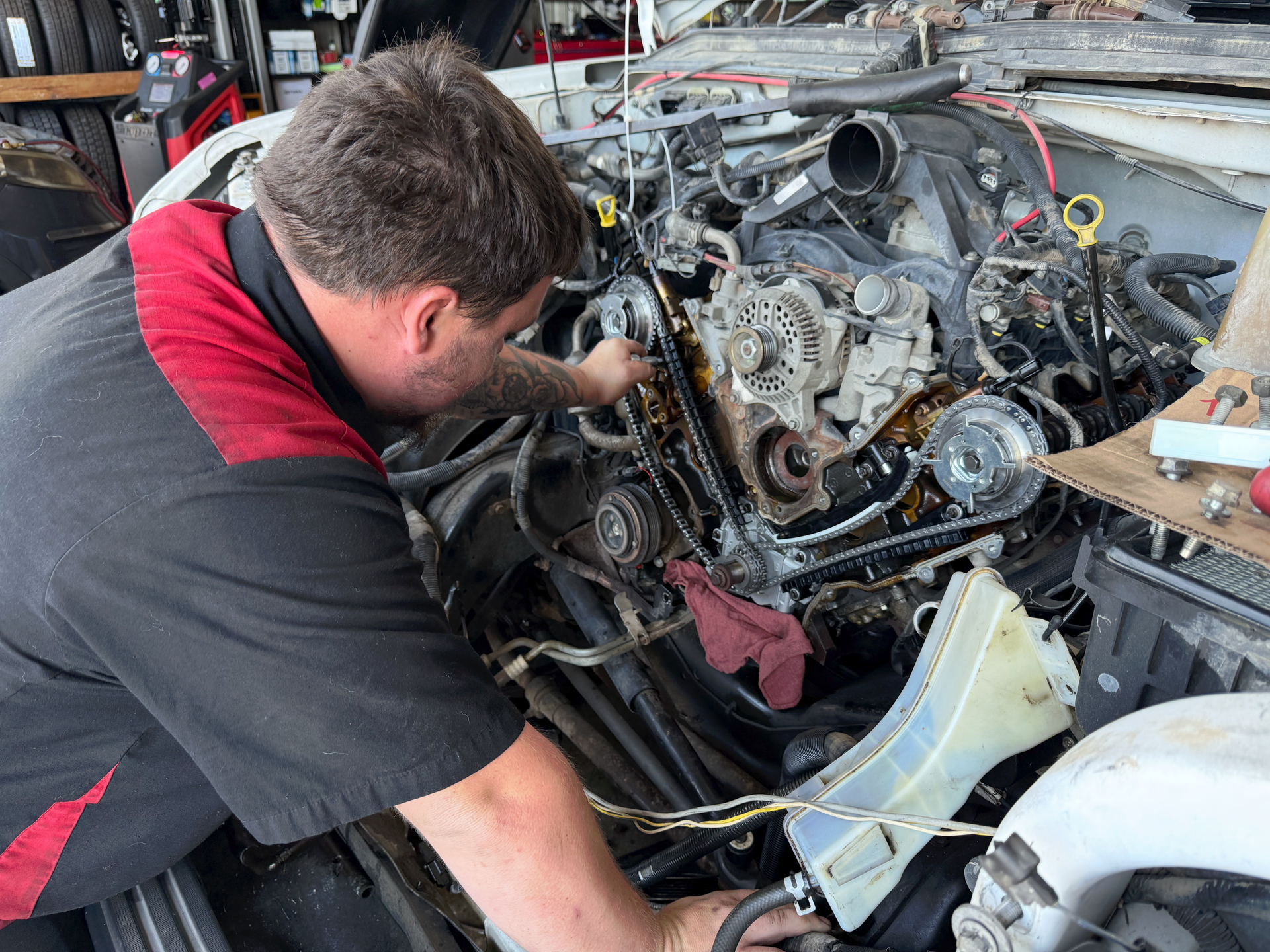 Mechanic working on a car engine. He's wearing a black and red shirt. Engine is open. Tools are on a small tray. | Auto Masters Repair Shop