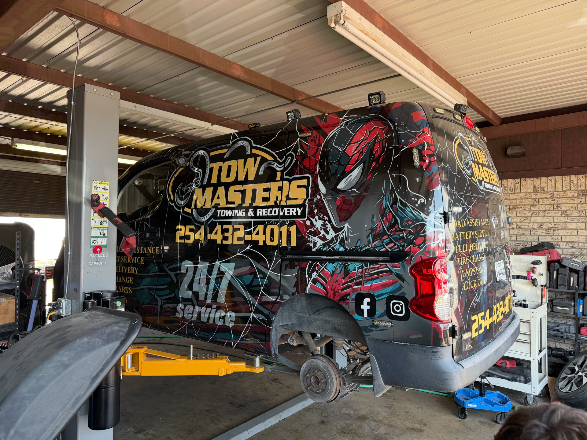 Tow Masters tow truck raised on a lift in a shop. Black and red graphics, missing rear wheel. | Auto Masters Repair Shop