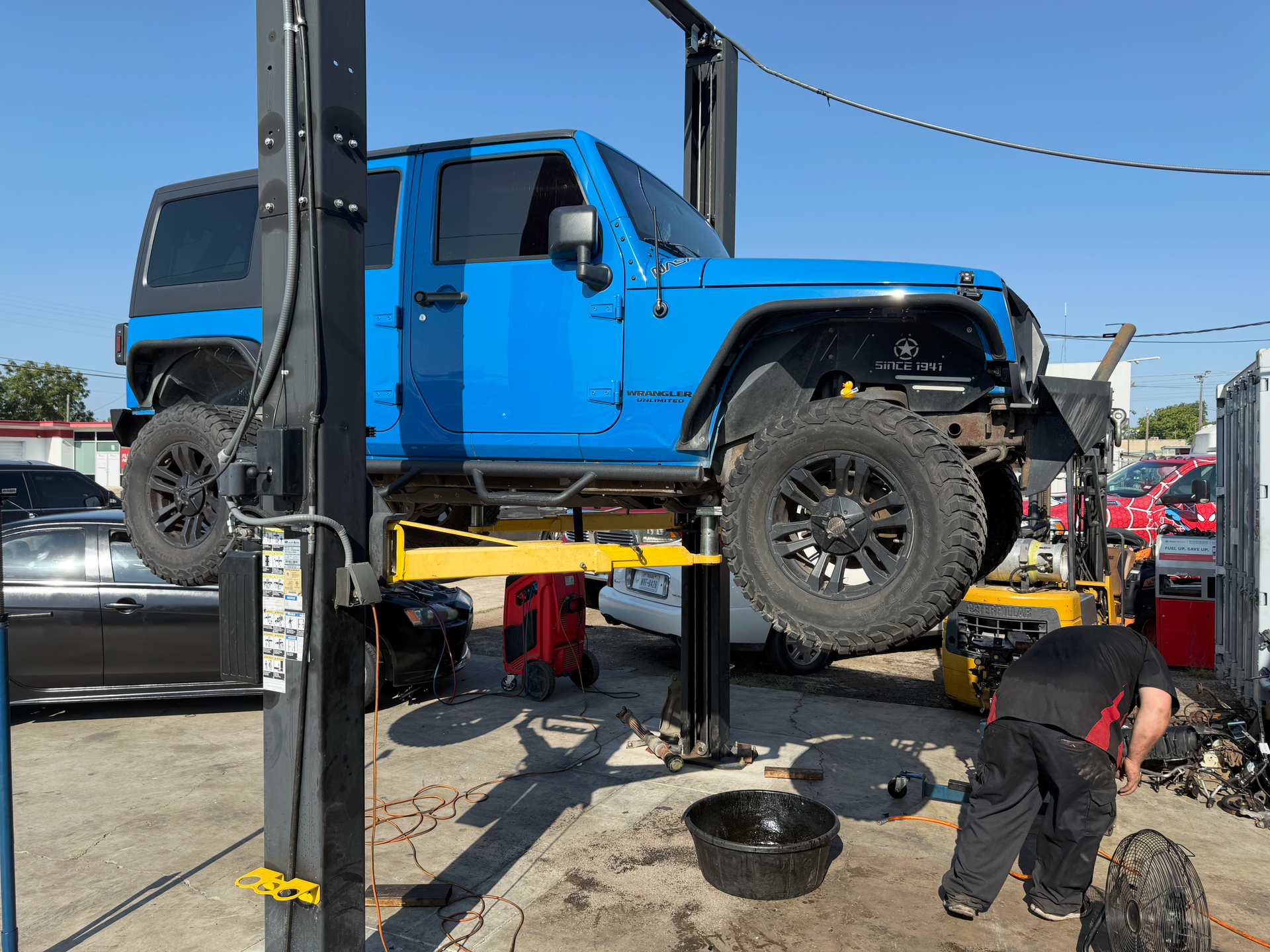 Blue Jeep Wrangler on a lift, being worked on by a mechanic outside. | Auto Masters Repair Shop