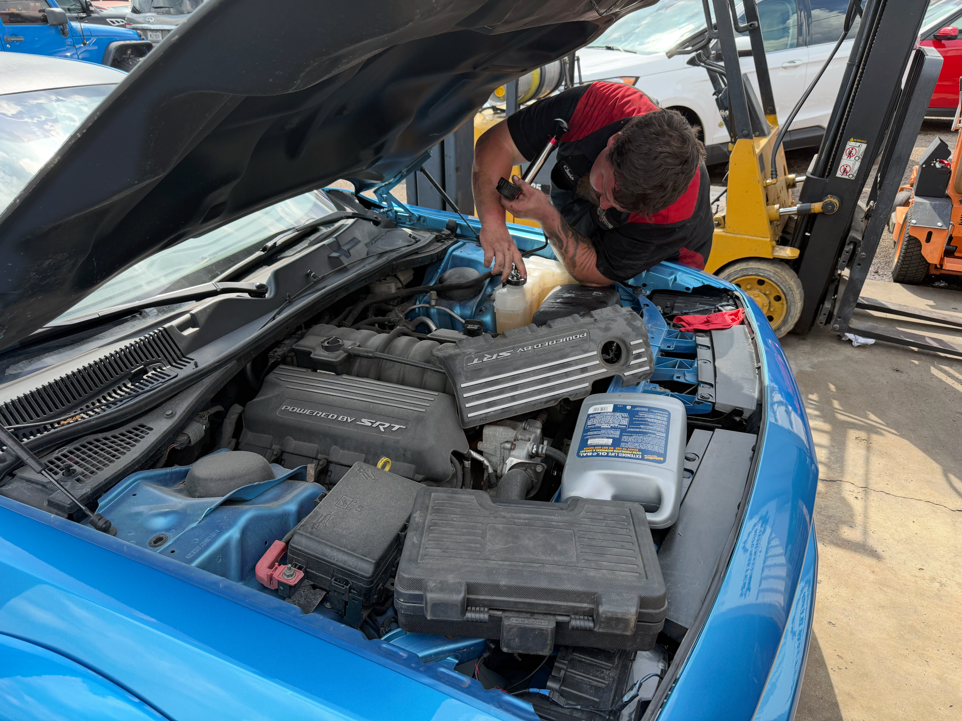 Mechanic working on a blue car engine with a container of oil, outside, next to a forklift. | Auto Masters Repair Shop