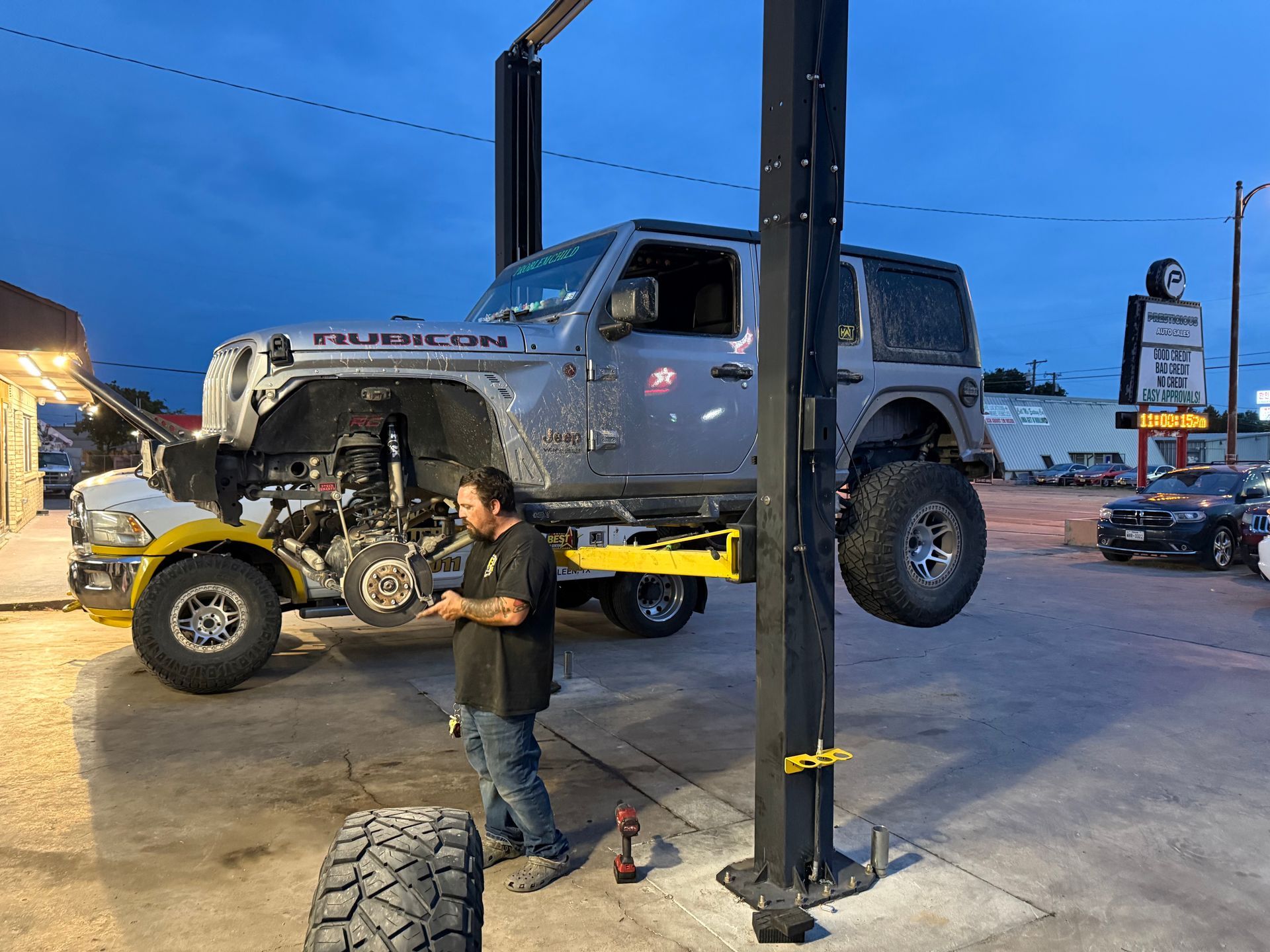 Mechanic working on lifted Jeep Wrangler Rubicon. Outside, under lift, at dusk. | Auto Masters Repair Shop