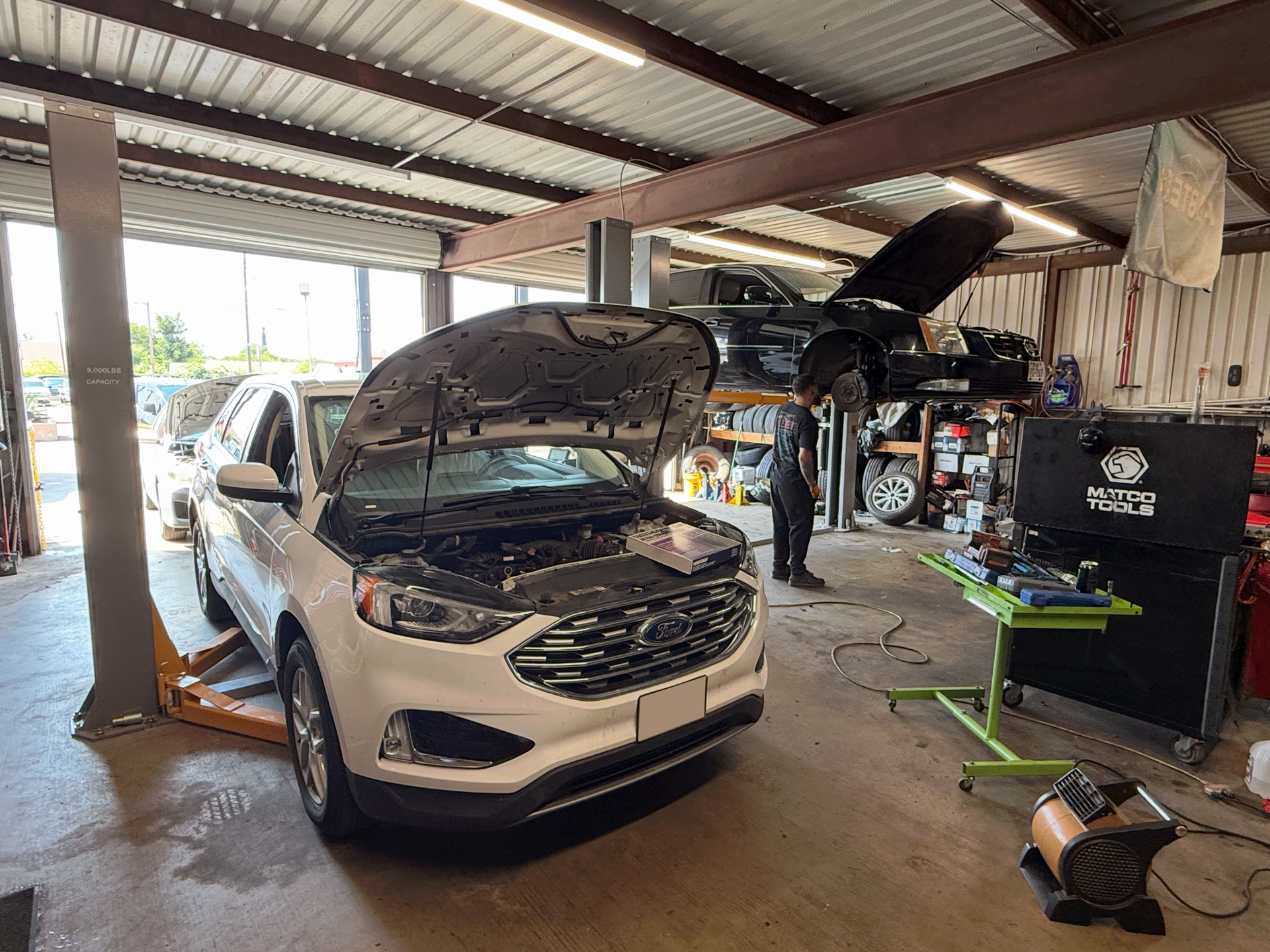 White Ford Edge with open hood in auto shop; black truck on lift in background. | Auto Masters Repair Shop
