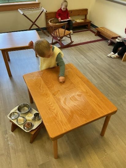 Child touches tabletop, small tray of metal dishes nearby, other children read in background.