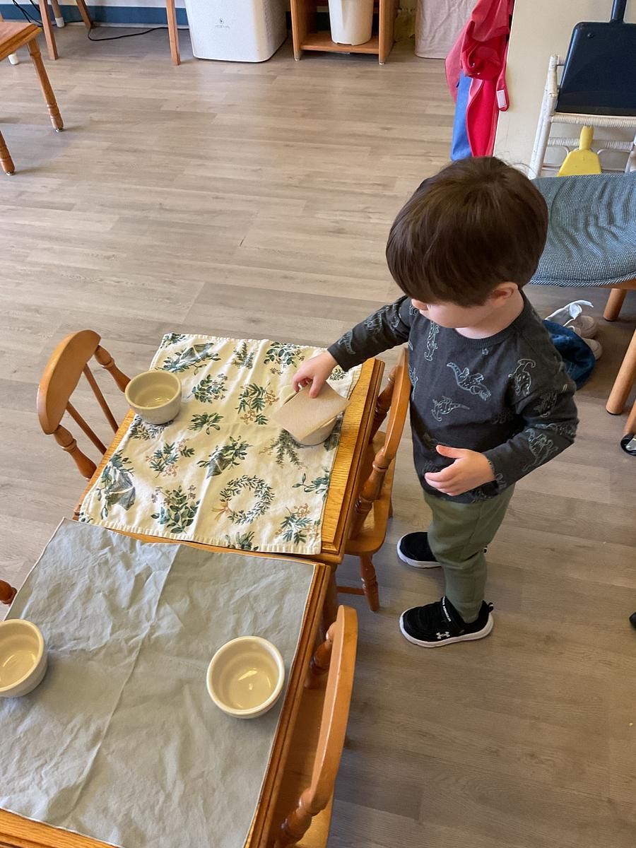 A child stands near a small table, placing a small board on the table. There are bowls on the table.