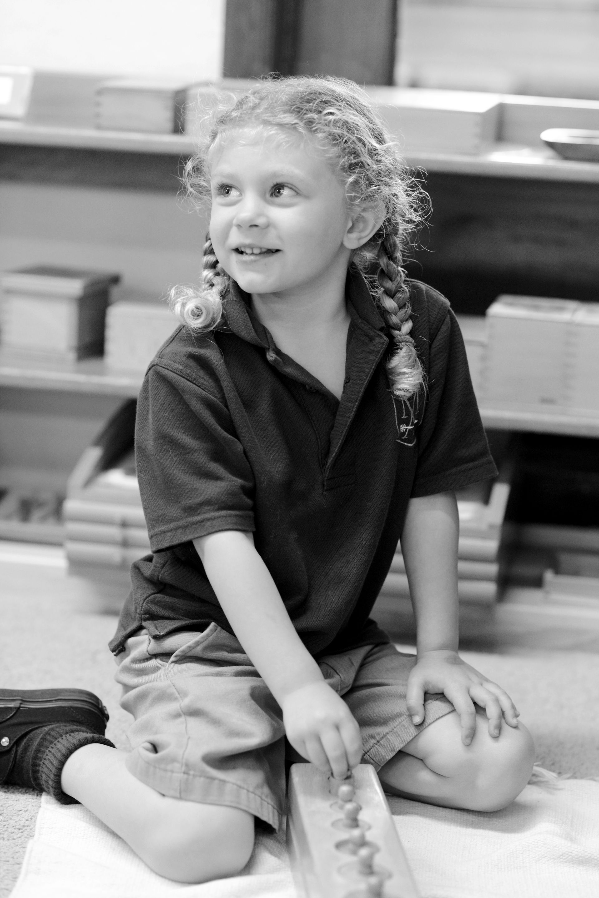 A young girl with braided hair smiling while working with a Montessori cylinder block in a classroom.