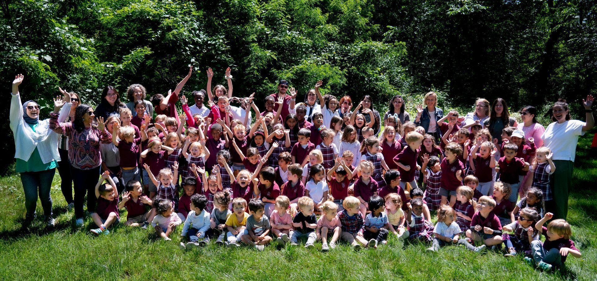 Group photo of Montessori Academy of New Jersey students and staff for the 2023-2024 school year, taken outdoors with everyone waving and smiling in a lush green setting.