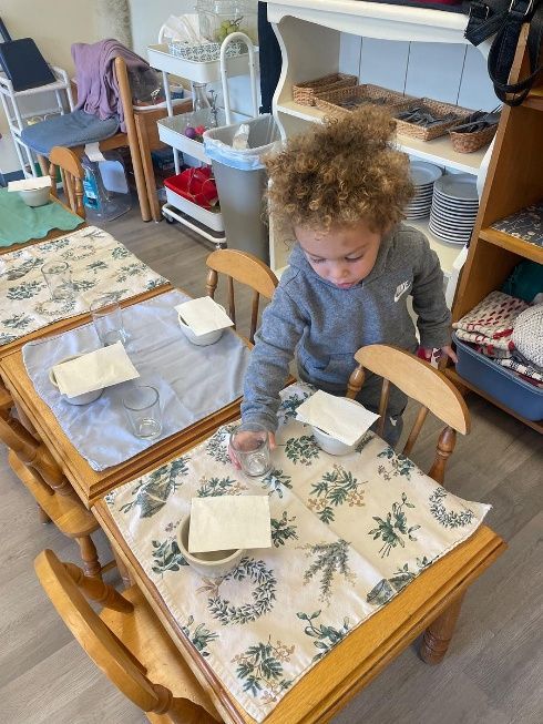 Young child at a table with cloth placemats, reaching for a bowl. Shelves and chairs are visible.