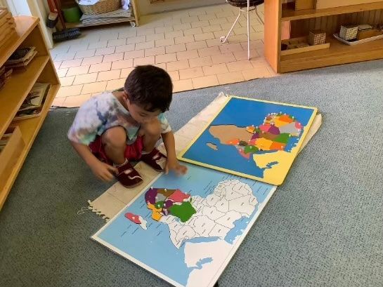 Child kneeling on a rug, working with colorful puzzle maps in a classroom.