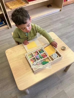 Child in a green sweater seated at a small table, using a puzzle tray in a classroom setting.