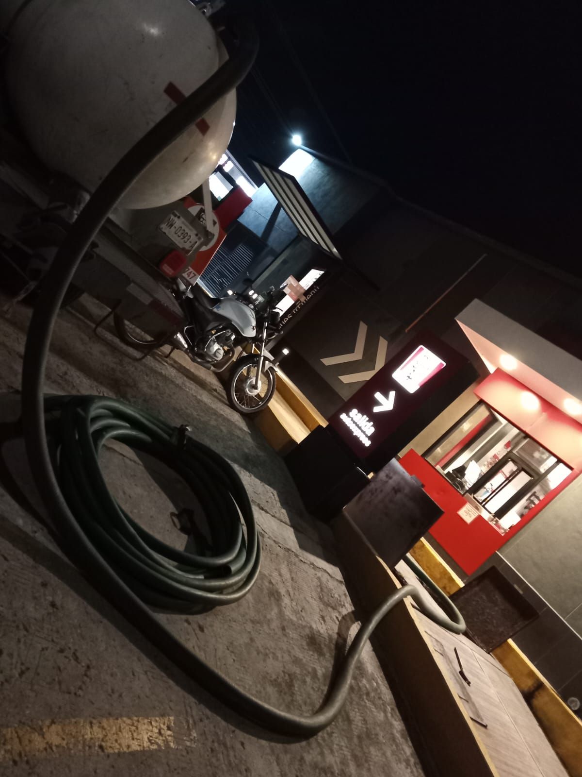 Escena oscura y nocturna: una motocicleta cerca de un drive-through, una manguera enrollada en el suelo y un tanque blanco sobre su cabeza.