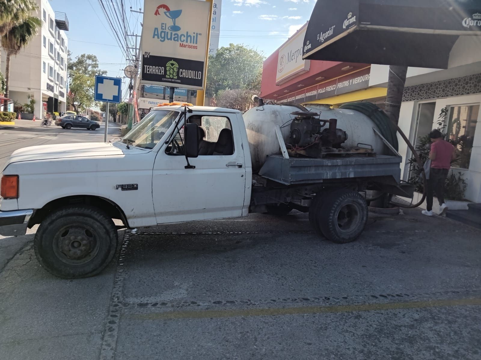 Un camión blanco con cisterna está estacionado, posiblemente bombeando, cerca de un edificio. Hay gente cerca.
