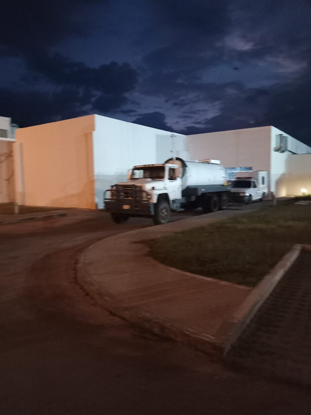 Camión de agua blanca estacionado frente a un edificio al anochecer, cielo nublado oscuro.