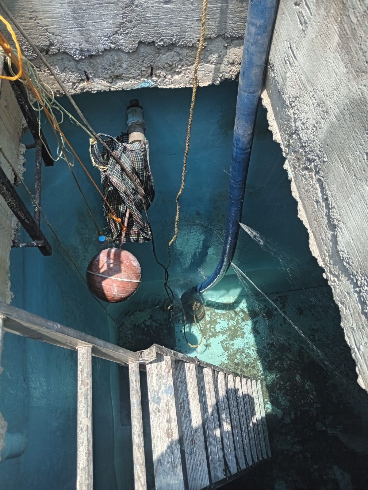 Mirando hacia abajo a una cisterna profunda llena de agua con tuberías, una escalera y un objeto colgante, posiblemente una bomba.