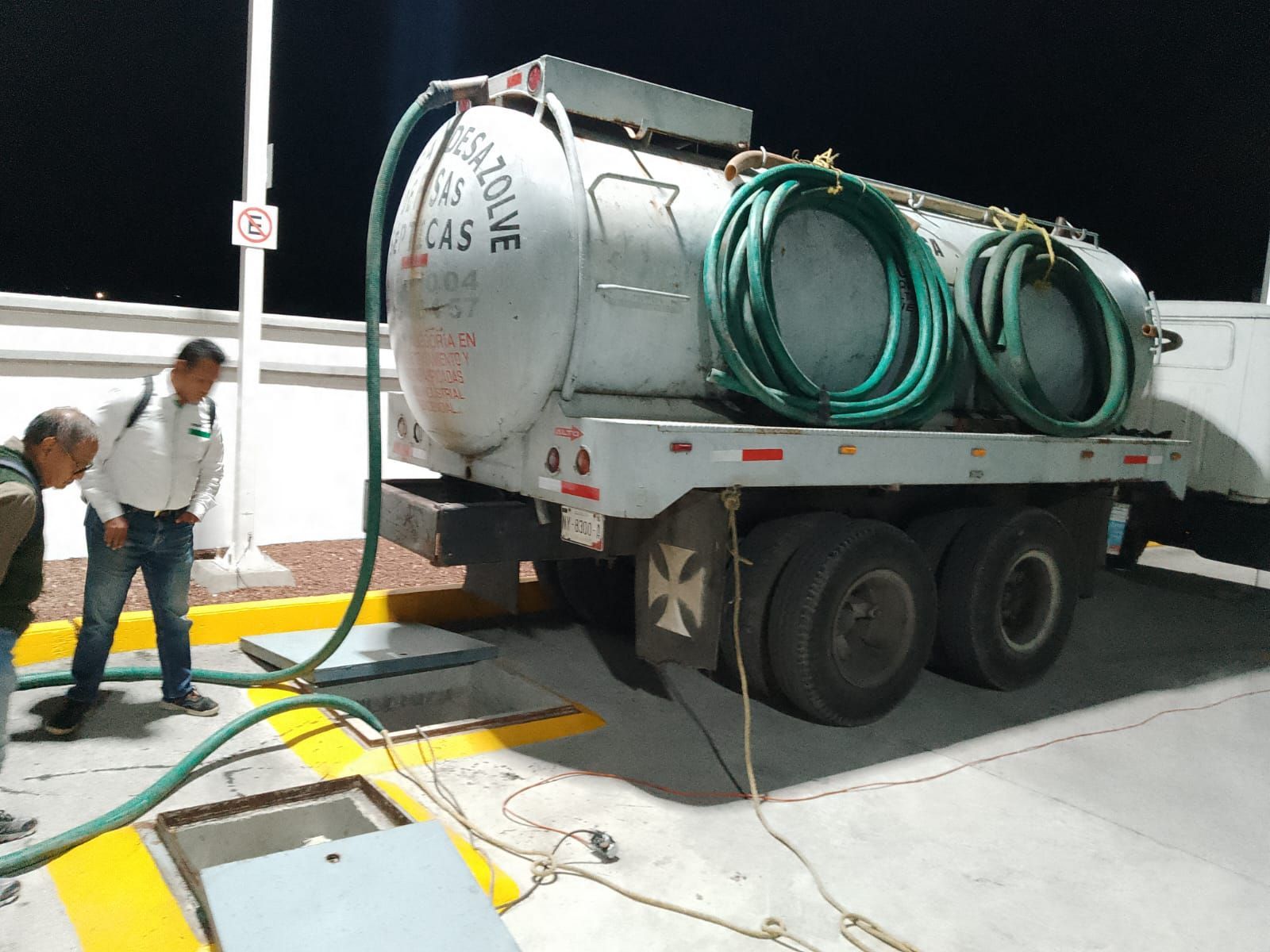Dos hombres cerca de un camión cisterna de gas, con las mangueras conectadas, de noche. El tanque dice