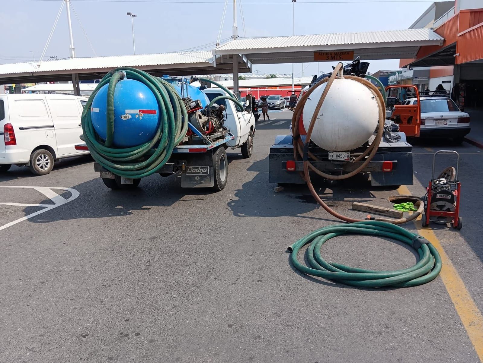 Dos camiones con cisternas y mangueras estacionados en un estacionamiento. Cisternas azules y blancas, pavimento gris.