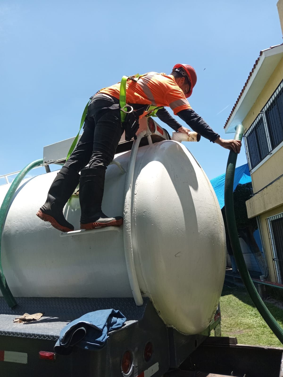 Trabajador con arnés de seguridad sobre un tanque blanco, conectando una manguera. Cielo azul al fondo.