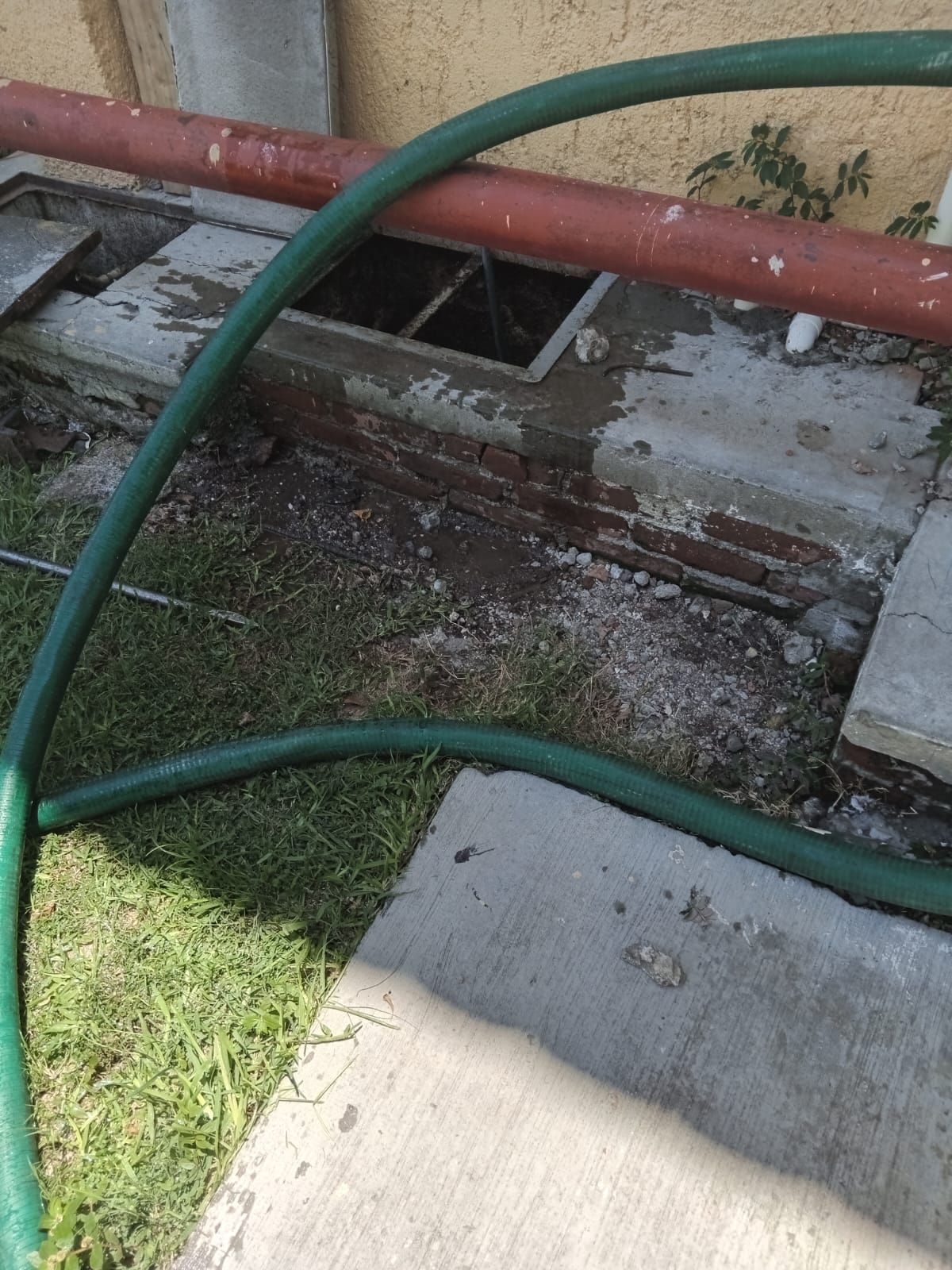 Manguera de jardín verde cerca de una estructura de ladrillo con escotillas abiertas y tubería roja.