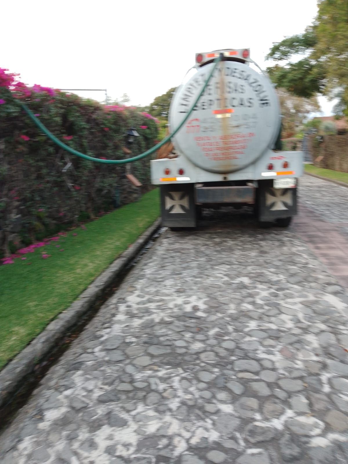 Camión tanque séptico en camino de entrada adoquinado, manguera conectada a un seto verde, frente a una zona residencial.