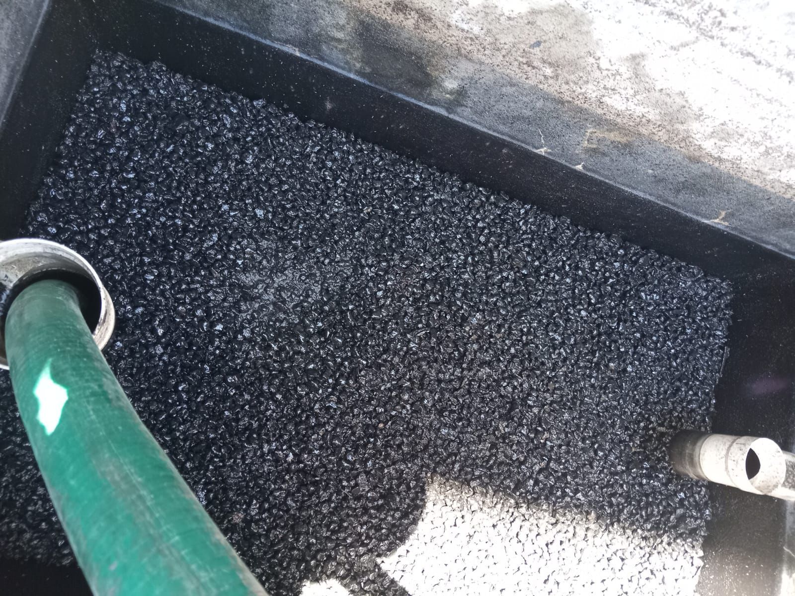 Material granular negro en un recipiente rectangular, una manguera verde rocía agua.